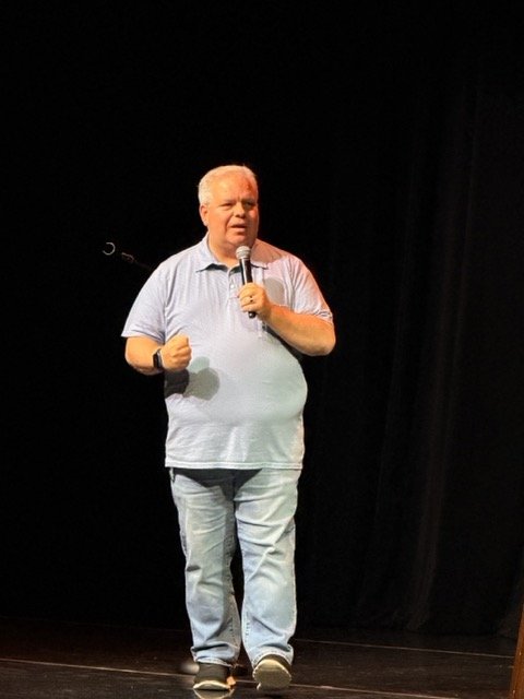A man with gray hair, wearing a light blue polo shirt and light blue jeans, is speaking into a microphone on stage with a black background.