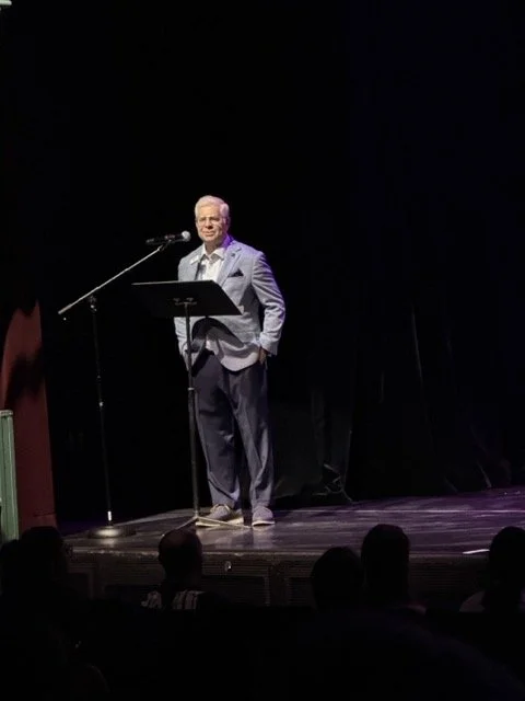 A man in a gray blazer and dark pants stands on a stage behind a podium, speaking into a microphone, with a black curtain backdrop.