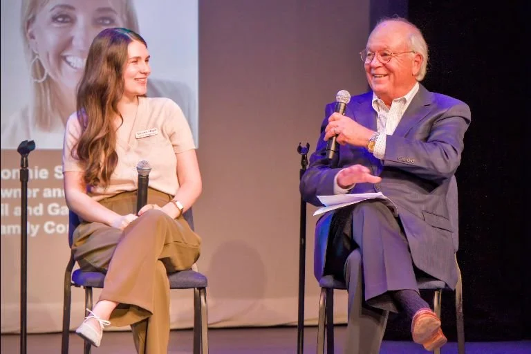 A young woman and an older man sit on stage in conversation, both holding microphones and smiling. The woman is wearing a beige top and brown pants, while the man is in a gray suit and glasses. A large screen in the background displays the woman's fa