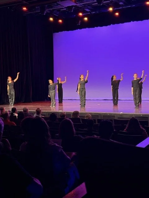 Six dancers in black and gray outfits perform on stage with purple background, audience watching in theater.
