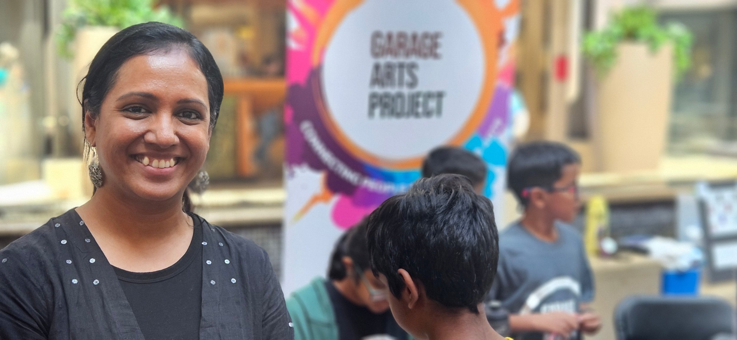 Woman smiling next to a 'Garage Arts Project' sign, with children in the background participating in a workshop or activity.