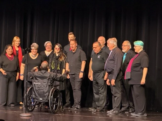 Group of ten people standing on stage, being recognized or awarded, with one person in a wheelchair facing them, in a theater or auditorium setting.