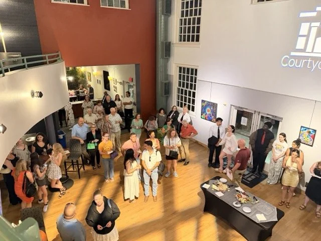 People gathering in a spacious art gallery with wooden floors, white and orange walls, and colorful artwork on display.