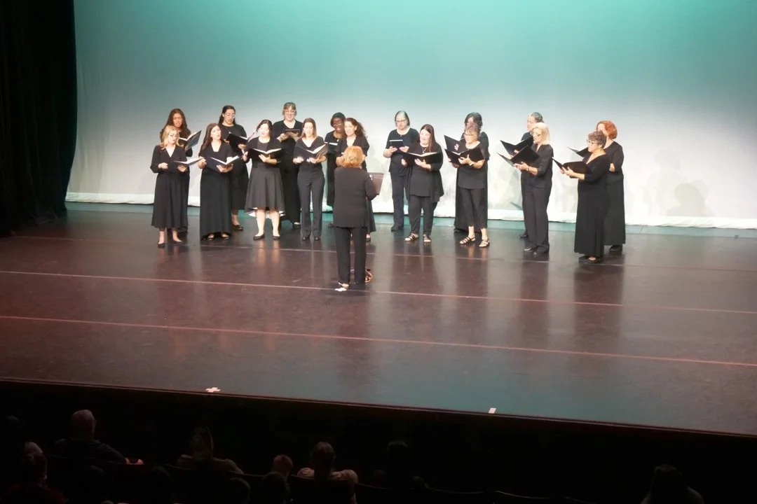 Chorus of women singing on stage with conductor leading.