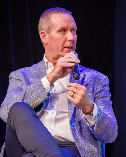 A man with short hair, wearing a light blue blazer and white shirt, holding a microphone and sitting on a chair on stage.