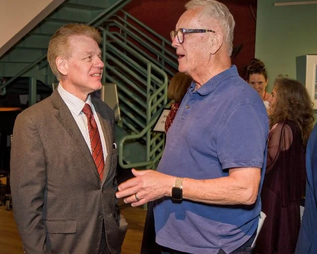 Two men are engaged in conversation at an indoor social event, with other people in the background.
