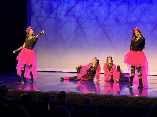 Four young girls performing a dance on stage in pink skirts and black tops, with two standing and two sitting or lying on the floor, in front of an audience.