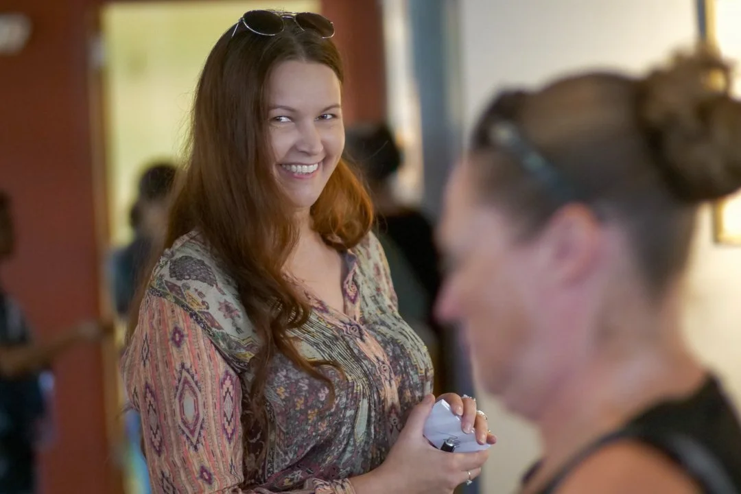 A woman with long red hair smiling at another woman with glasses in an indoor setting.