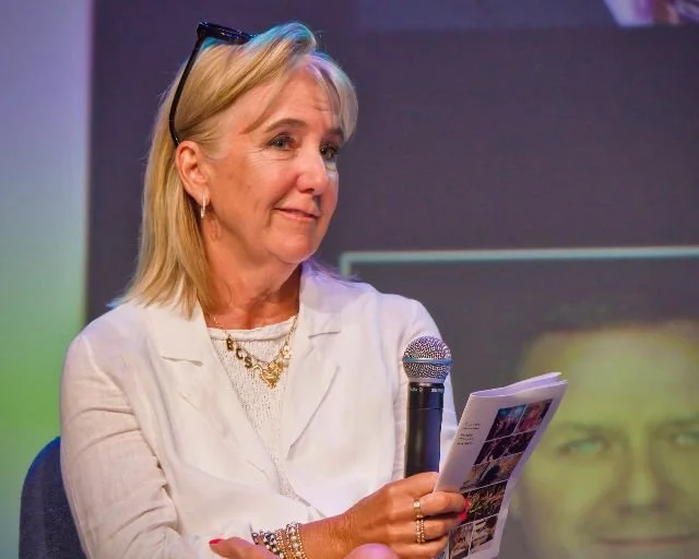An older woman with blonde hair, wearing glasses on her head, a white blazer and gold jewelry, holding a microphone and a pamphlet, sitting in front of a dark background, possibly during a presentation or event.