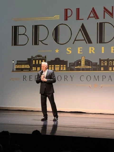 A man in a suit holding a microphone standing on a stage in front of a large screen that reads 'Plan Broad Series' and 'Reedory Company' with illustrations of buildings and a star.