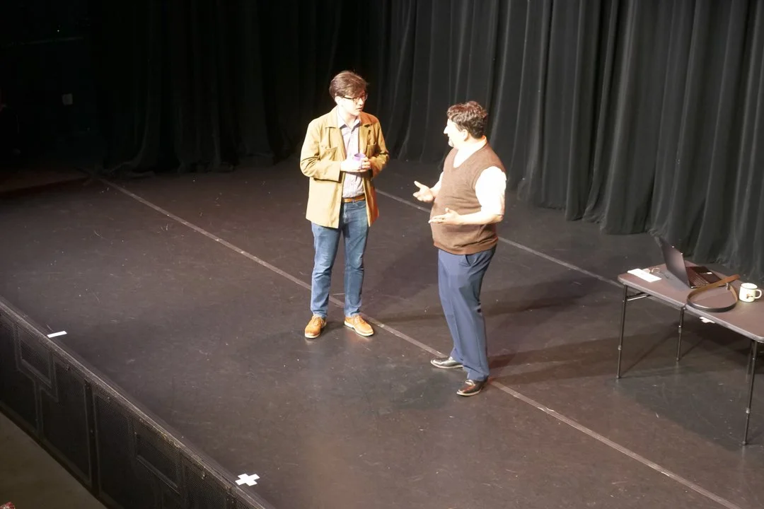 Two men are talking on a stage with black curtains behind them. One man is holding a pink mug, and there is a table with a laptop, a phone, a notebook, a cup, and some papers on it.