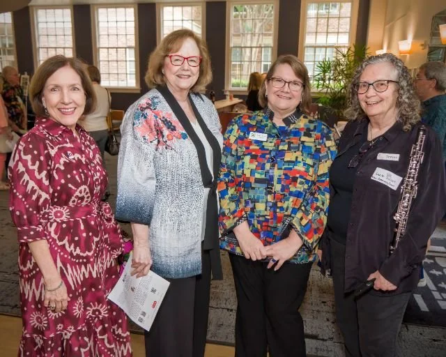 Four women standing together at an indoor event, smiling, with other people in the background and large windows letting in natural light.