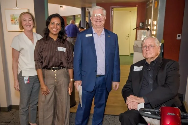Four people, three standing and one sitting, at an indoor event, smiling for the photo. All are wearing professional attire and name tags.