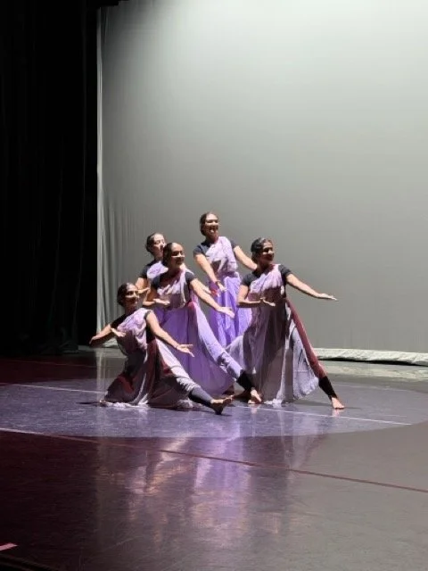 Five young girls in matching purple dresses performing a dance on stage.