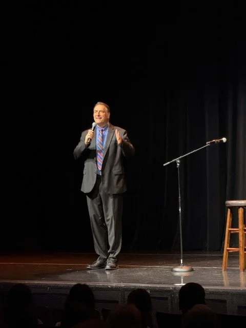 Man in a suit performing stand-up comedy on stage with a microphone, a stool, and a black curtain backdrop.