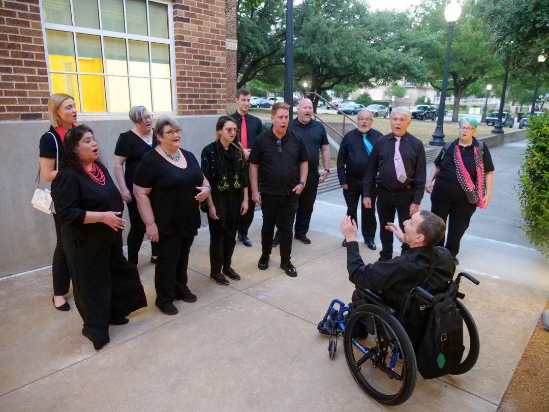 A choir of eleven people singing outdoors, facing a conductor in a wheelchair, in front of a brick building and trees.