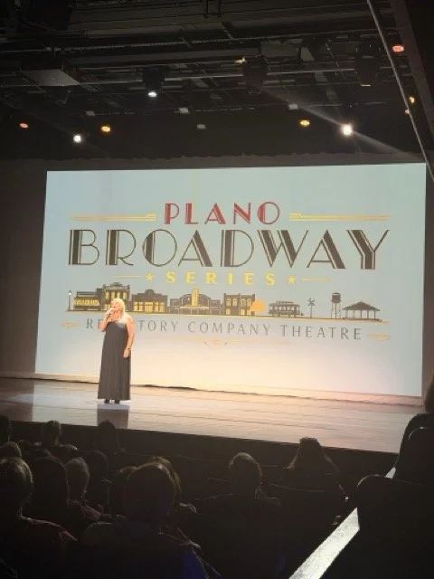 A woman standing on a stage in front of a large screen displaying the Plano Broadway Series logo, with an audience seated in front.