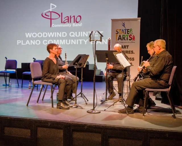 Group of five musicians seated on stage playing wind instruments, with music stands and sheet music in front of them, during a performance at the Plano Community Band event, with a backdrop displaying the event name and a colorful banner promoting ar