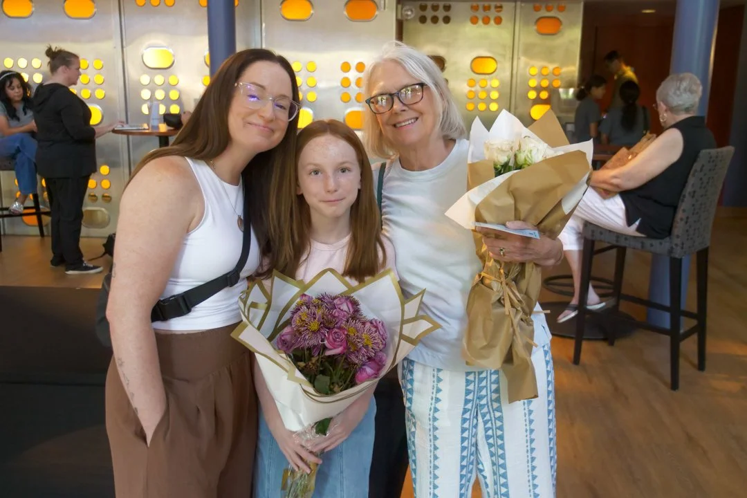 Three women and a girl with flowers posing together in a modern indoor setting, with other people in the background.