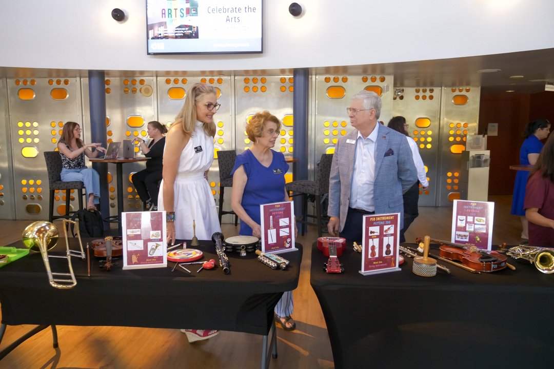 People at a table displaying small musical instruments and accessories in an indoor setting, with a few individuals using laptops in the background.