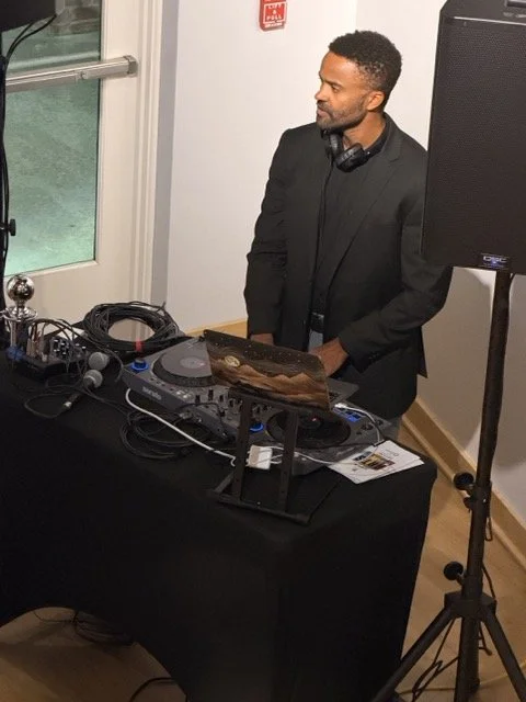 A man in a black suit standing behind a DJ setup with turntables, a mixer, headphones around his neck, and a speaker on a stand in a room with a door and a window.