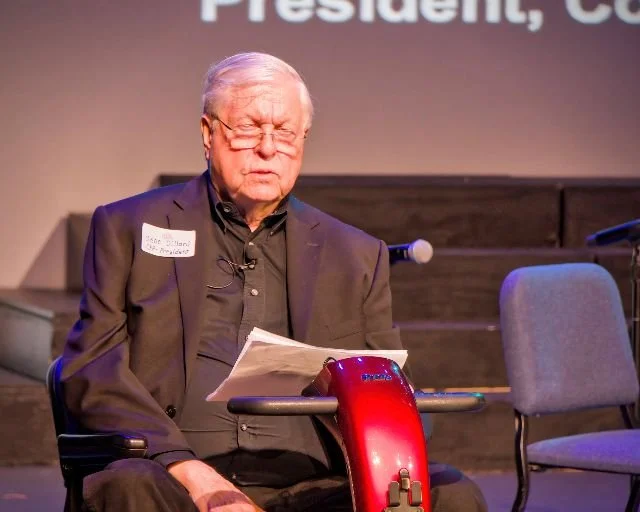 An elderly man with glasses, dressed in a dark suit, sitting in a wheelchair, holding papers and appearing to squint or be upset at an event.