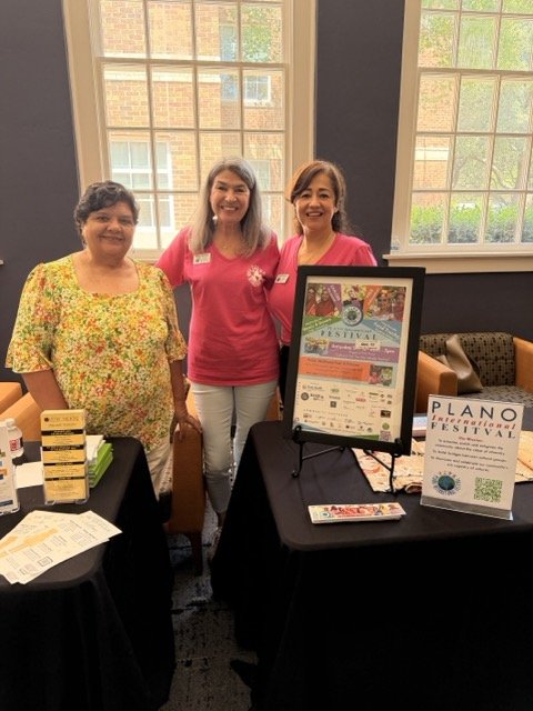 Three women standing behind promotional tables at an indoor event, with large windows in the background. One woman is wearing a floral blouse, and the other two are in pink shirts. The tables display flyers, brochures, and a framed poster for the Pla