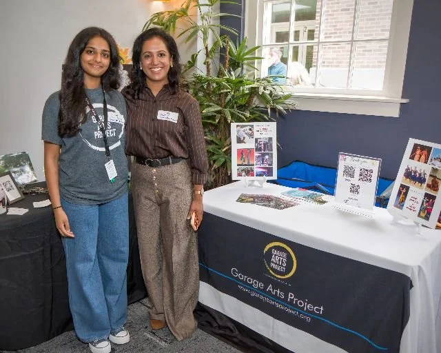 Two women standing indoors behind a display table at an event, with the table featuring a black tablecloth with the Garage Arts Project logo and website URL. The table has various informational materials, and there are frames with photos on the table