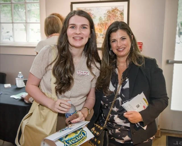 Two women smiling at a photo event, one with long dark hair wearing a beige shirt, the other with dark hair in a black blazer, holding a booklet or program.