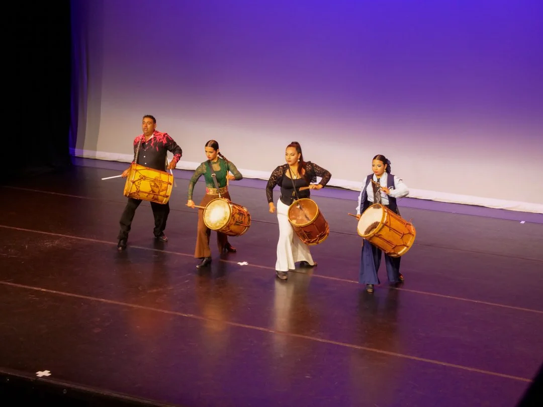 Four performers, one man and three women, playing drums on stage during a dance performance with a purple-hued backdrop.
