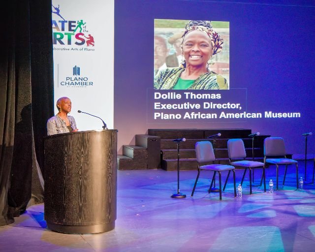 A woman stands at a podium on a stage, giving a speech or presentation. Behind her is a large screen displaying a photo of another woman and text that reads 'Dollie Thomas, Executive Director, Plano African American Museum.' There are four empty chai