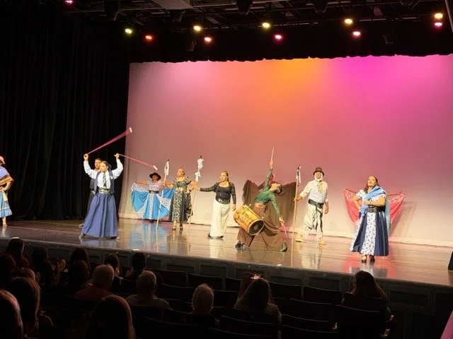 Theater stage performance with actors in period costumes, holding props like staffs, drums, and fabrics, with a colorful backdrop and audience watching.