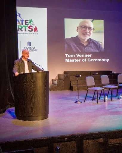 A man standing at a podium on a stage giving a speech. Behind him is a large screen displaying a photo of Tom Venner with the text 'Tom Venner Master of Ceremony.' To his right, there are four empty chairs and three microphone stands on the stage. Th