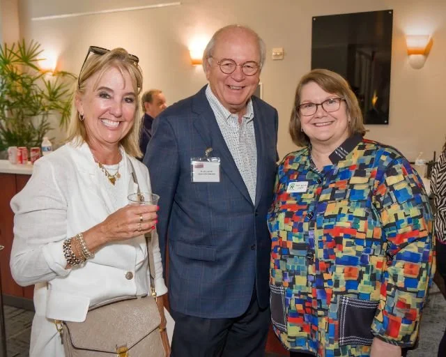 Three people smiling at a social event indoors, two women and one man, standing close together. The woman on the left is holding a drink, wearing a white jacket over a beige top, and has glasses on her head. The man in the middle is dressed in a dark