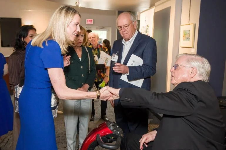 A woman in a blue dress shaking hands with a seated older man in a black suit at an art gallery event, with several others observing in the background.