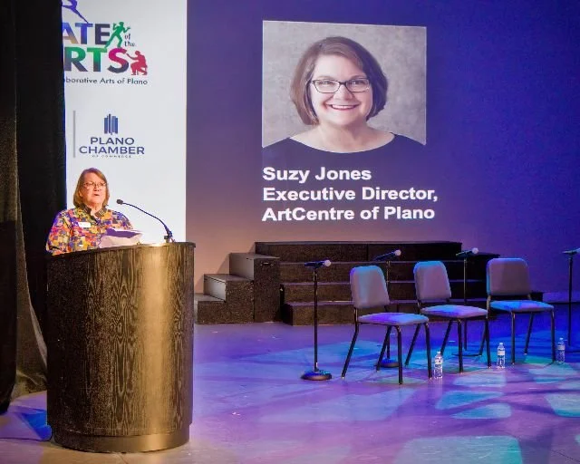Woman speaking at a podium during an event with a large screen displaying her photo and title, 'Suzy Jones, Executive Director, ArtCentre of Plano.'