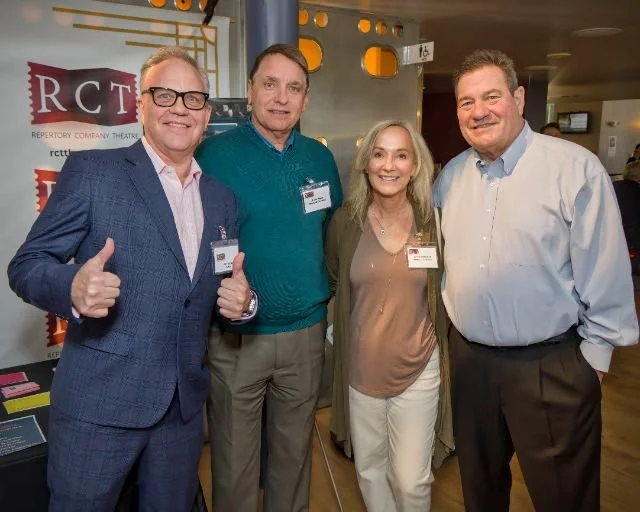 Four people standing together indoors, posing for a photo during a professional event. They are smiling and wearing business casual attire with name tags. The background includes a banner with red and white text and a theater-themed decor.