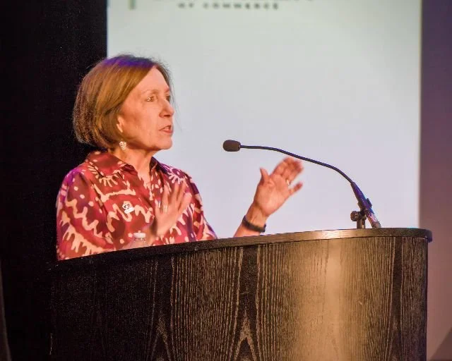 A woman with short brown hair and wearing a red patterned shirt delivers a speech at a podium with a microphone.