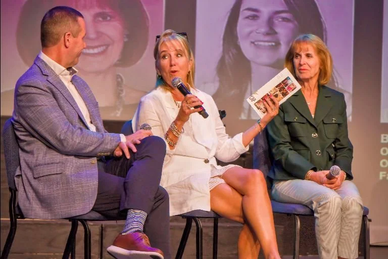 Three people sitting on stage during a panel discussion, with a woman in the middle speaking into a microphone and holding a pamphlet, behind a backdrop displaying large photos of faces.