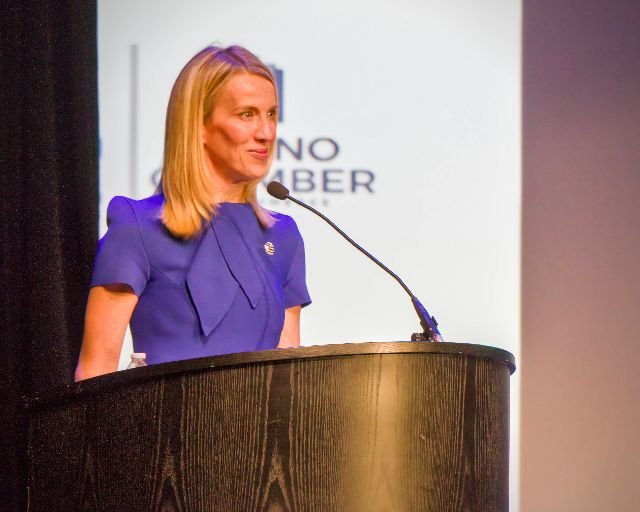 A woman with shoulder-length blonde hair speaking at a podium with a microphone during a presentation or conference.