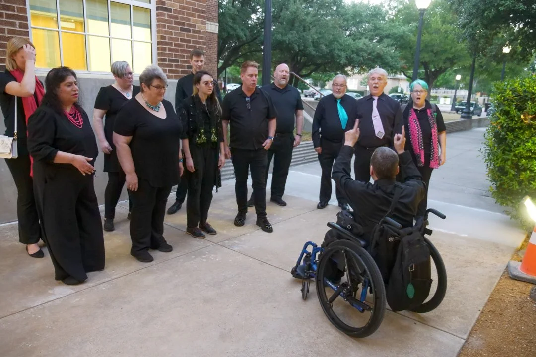 A group of people singing outdoors, facing a person in a wheelchair who is conducting them.