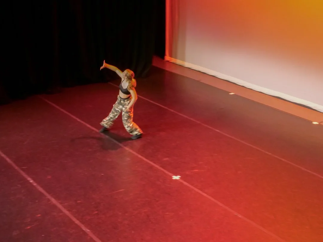 A person dancing on a stage with a dark curtain behind them and a white screen to the right, wearing camouflage pants and a black top.