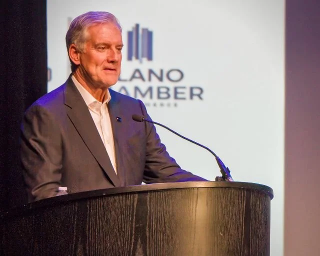 Older man in a gray suit speaking at a podium during a presentation or conference. Background features a white screen with partial logo and text.