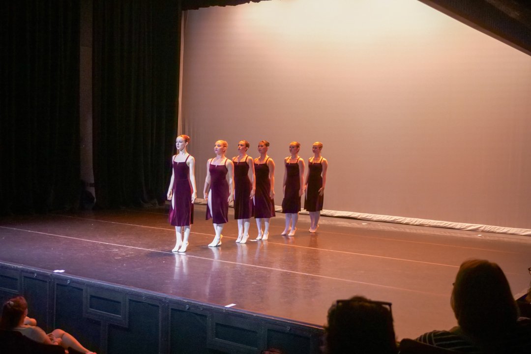 Six ballet dancers in purple costumes perform on stage with a plain backdrop while audience members watch.