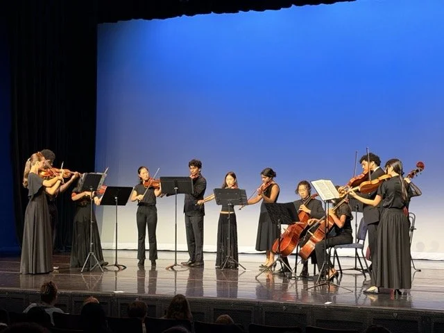 A group of nine musicians in black clothing performing on a stage with blue background, playing string instruments including violins, viola, cello, and double bass.