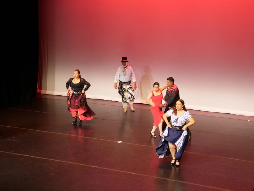 Group of five performers on stage, dressed in colorful costumes, dancing with a plain white backdrop and pinkish lighting.