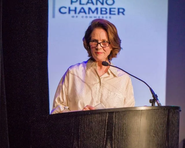 A woman with short, wavy brown hair and glasses standing behind a black podium, speaking into a microphone, with a screen in the background displaying 'Plano Chamber'.