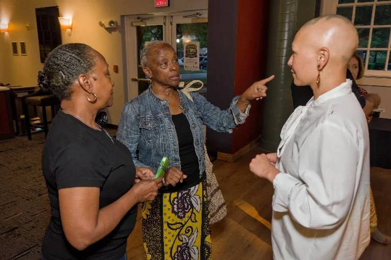 Three women engaged in a conversation indoors, with one woman pointing her finger at another woman, who has a shaved head and is wearing a white jacket.