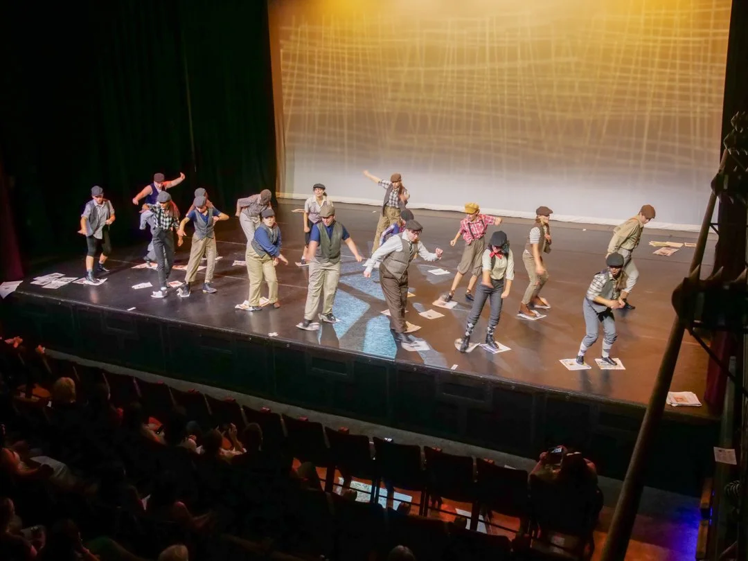 A group of children performing a dance on stage in front of an audience, dressed in retro outfits with hats and suspenders, surrounded by scattered papers.