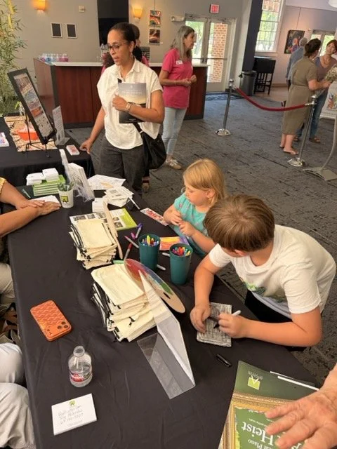 Children and adults at an indoor event registration table with papers, pens, and a water bottle.